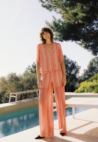 Femme souriante debout près de la piscine, portant un haut à manches courtes et un pantalon à jambes larges assortis orange et blanc à motifs, en plein air.