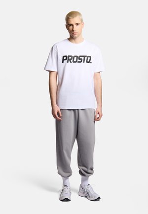 Młody mężczyzna stojący, ubrany w białą koszulkę "PROSTO.", szare spodnie dresowe, białe skarpetki i białe buty do biegania na jednolitym tle.