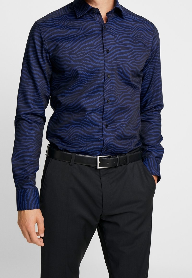 Knapp-upp skjorta i marinblått tyg med en texturerad vågmönster i mörkare nyanser. Långa ärmar, krage och figurnära design. Bärs med svart bälte och byxor.