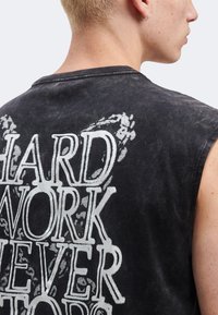 Νεαρό άτομο με κοντά ξανθά μαλλιά φοράει μαύρο αμάνικο τοπ με λευκό κείμενο στην πλάτη που γράφει «HARD WORK NEVER STOPS».