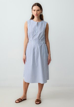 Robe sans manches rayée bleue et blanche avec un col rond, une taille cintrée et une jupe à hauteur de genoux. Portée avec des sandales plates marron.