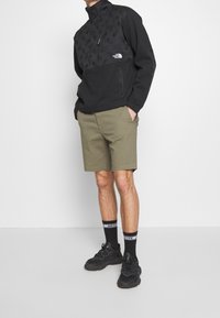 Svart huvtröja med mönstrad övre del, dragkedja vid kragen; olivgröna shorts; svarta sneakers och strumpor med vita detaljer.