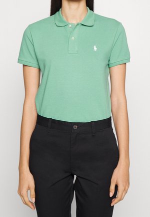 Persoon draagt een groen poloshirt met korte mouwen en een wit logo op de borst en zwarte high-waist broek tegen een effen achtergrond.