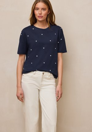 Vrouw draagt marineblauw T-shirt met verspreide witte hartjesprint en gebroken witte broek, staand voor een effen beige achtergrond.