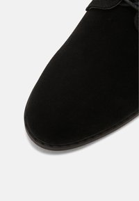 Bout de chaussure en daim noir avec couture visible et devant arrondi sur un fond clair.