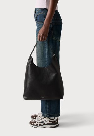 Black leather hobo bag with a curved top opening and a flat base, featuring a subtle logo. Paired with distressed blue jeans and white sneakers.
