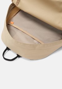 Interno dello zaino beige, caratterizzato da una fodera in tessuto liscio, una tasca in rete e spallacci neri a contrasto per un facile trasporto.