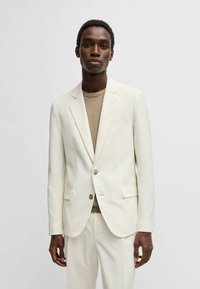 Blazer bianco con texture, colletto a forma di notched, chiusura con un bottone e tasche frontali. Indossato sopra una camicia beige, abbinato a pantaloni bianchi coordinati.