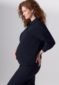 Fekete kismama pulóver gallérral, laza szabású, hosszú ujjú, texturált anyagból. Fekete, hozzá illő nadrággal párosítva.