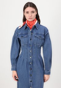Modra denim obleka s dolgimi rokavi in gumbi, žepoma na prsih in oblikovano pasovno zasnovo. Opremljena z rdečo šalom z vzorci.