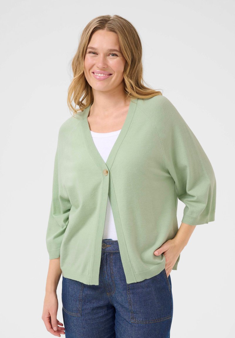 Cardigan vert clair en tissu doux, avec un col en V, des manches trois-quarts et une fermeture par un unique bouton en bois. Associé à un haut blanc.