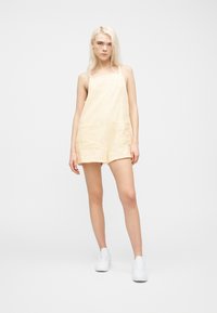 Νεαρή γυναίκα με μακριά ξανθά μαλλιά, ντυμένη με μπεζ ολόσωμο σορτς χωρίς μανίκια με υφή και λευκά αθλητικά παπούτσια, στέκεται μπροστά σε απλό λευκό φόντο.