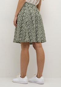 Jupe verte à motif avec une taille élastique, arrivant au genou, présentant des designs abstraits blancs. Elle est associée à des baskets blanches.
