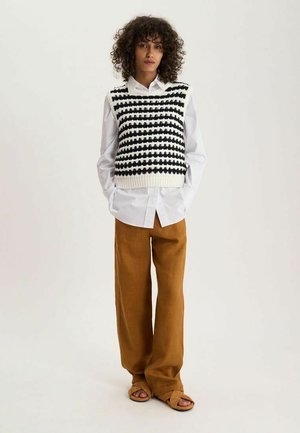 Femme aux cheveux bouclés portant une chemise blanche, un gilet tricoté rayé noir et blanc, un pantalon large marron et des sandales marron, debout les mains dans les poches.