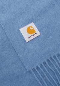 Svetlomodrý vlnený šál so štítkom so žltým logom. Šál má strapcové konce a mäkký, textúrovaný povrch.