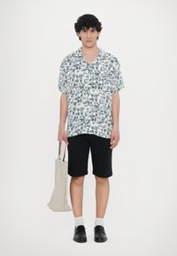 Λευκό πουκάμισο κοντομάνικο με σκούρο πράσινο φυτικό σχέδιο, συνδυασμένο με μαύρες βερμούδες. Κρατάει μια μπεζ τσάντα, φοράει σκούρα παπούτσια και λευκές κάλτσες.