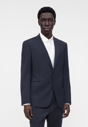 Jonge man die een donkerblauw colbert en broek draagt met een wit overhemd met knopen, staand tegen een effen lichte achtergrond.