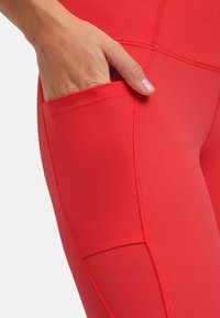 Leggings sportivi rossi realizzati in tessuto elasticizzato, con una tasca laterale e cuciture piatte per il comfort. Il design è aderente e affusolato.