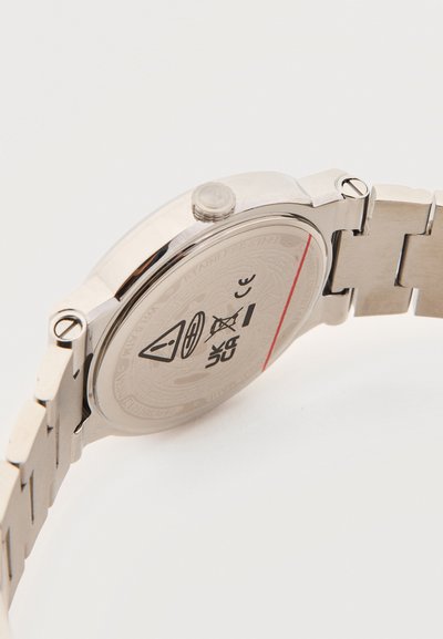 Μεταλλικό ρολόι χειρός γυρισμένο ώστε να φαίνεται η πίσω πλευρά με προειδοποιητικά σύμβολα και μεταλλικό μπρασελέ συνδεδεμένο σε λευκό φόντο.