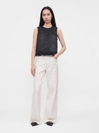 Femme aux longs cheveux foncés portant un haut noir sans manches et un pantalon blanc large, debout avec une main dans la poche sur fond blanc.