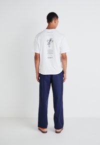 White cotton t-shirt with a graphic illustration on the back. Paired with navy blue wide-leg trousers and light brown wooden clogs.