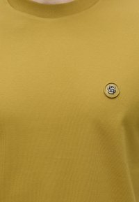 Μουσταρδί μπλούζα με στρογγυλή λαιμόκοψη και ένα μικρό στρογγυλό έμβλημα με ανάγλυφο ασημένιο αριθμό 8 στην αριστερή πλευρά στο στήθος.