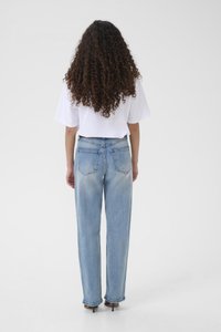 Pikkade lokkis juustega naine seisab seljaga vaataja poole, seljas valge lühikeste varrukatega särk, helesinised teksad ja pruunid kontsaga kingad valgel taustal.