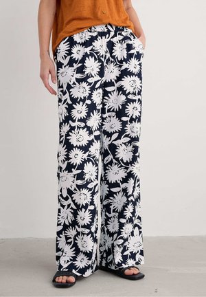 Person trägt lockere marineblaue Hosen mit großem weißem Blumenmuster, kombiniert mit schwarzen Sandalen und einem orangenen Oberteil. Hände in den Taschen.