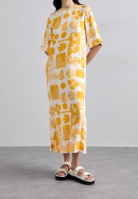 Robe jaune à motifs en tissu léger, présentant des dessins graphiques sur fond blanc. Associée à des sandales blanches épaisses.