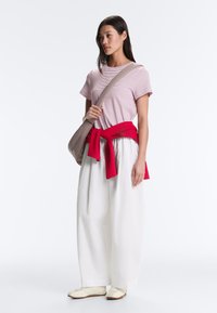 Femme portant un t-shirt rayé rose, un pantalon large blanc, des baskets blanches, un pull rouge noué à la taille et un sac bandoulière beige, debout regardant vers la gauche.