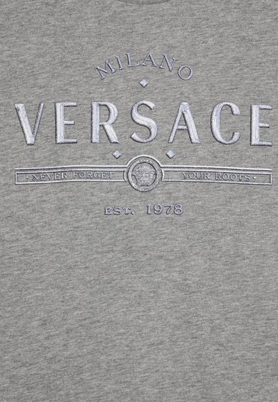 Camiseta de algodón gris con texto bordado en azul que dice "MILANO VERSACE" y la frase "NUNCA OLVIDES TUS RAÍCES", establecida en 1978.