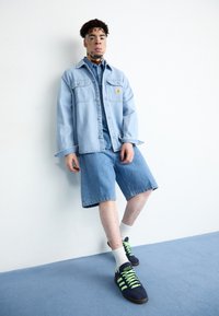 Ljusblå denimjacka över en denimskjorta, i kombination med ljusblå shorts. Svarta sneakers med neongröna snörningar och vita strumpor.