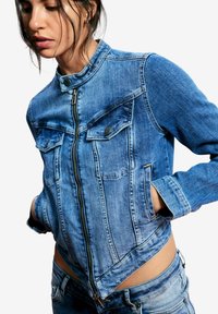 Hele sinises toonis teksajakk, millel on kõrge krae, eest tõmblukk, kaks rinnataskut ja lühendatud lõige kitsas siluetis.