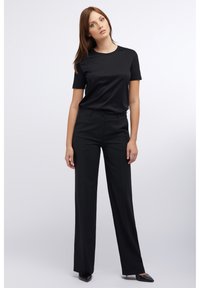 Schwarzes kurzärmeliges Hemd kombiniert mit hoch taillierter schwarzer Hose. Die Hose hat eine gerade Passform und eine subtile Textur; die Schuhe sind spitze schwarze High Heels.