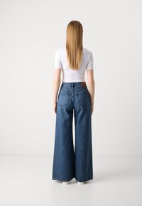 Jeans taille haute à jambes larges en denim bleu moyen, avec un design classique à cinq poches et des coutures contrastées. La vue arrière montre des cheveux longs.