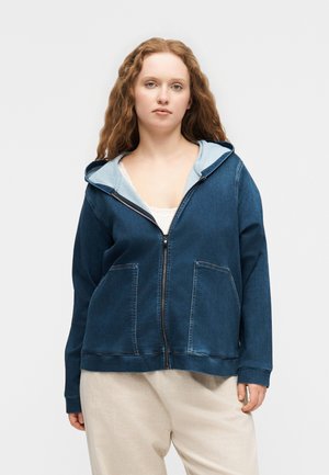 Junge Frau mit langen, lockigen Haaren, die einen dunkelblauen Hoodie mit Reißverschluss über einem weißen Top und beigen Hosen trägt, vor einem schlichten Hintergrund steht.