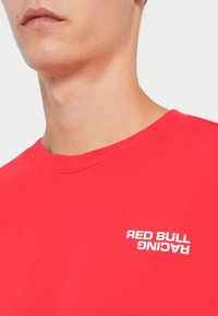 Piros pamut póló, kerek nyakkivágással; fehér "RED BULL RACING" felirattal a bal mellrészen. Sima textúra.