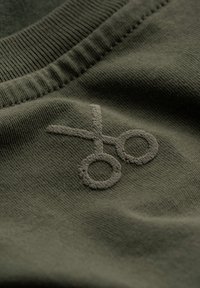 Tissu vert olive avec une texture lisse, présentant un logo de ciseaux en relief, créé avec un détail de couture légèrement surélevé.