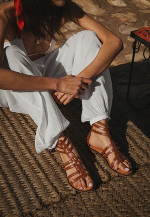 Kvinde, der sidder på et flettet tæppe iført hvide bukser, brune sandaler med stropper og et rødt tørklæde bundet i håret, med stenbelagt gulv og et lille bord i nærheden.