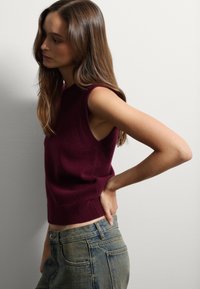 Jersey sin mangas burdeos con dobladillo acanalado y aberturas laterales, combinado con jeans de mezclilla rasgados de cintura alta.