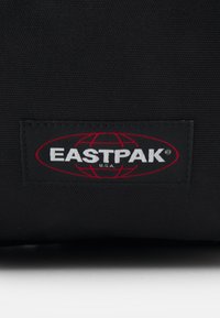 Černá textilní taška s výraznou oválnou nálepkou. Nálepka obsahuje bílé písmo "EASTPAK" a červený design glóbu. Texturovaný povrch.