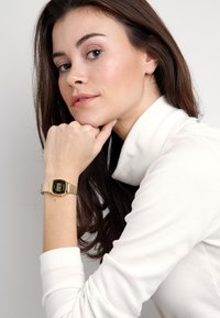 Goldene Digitaluhr mit rechteckigem Ziffernblatt und Gliederband aus Metall, am Handgelenk getragen. Der Hintergrund ist schlicht und hell.