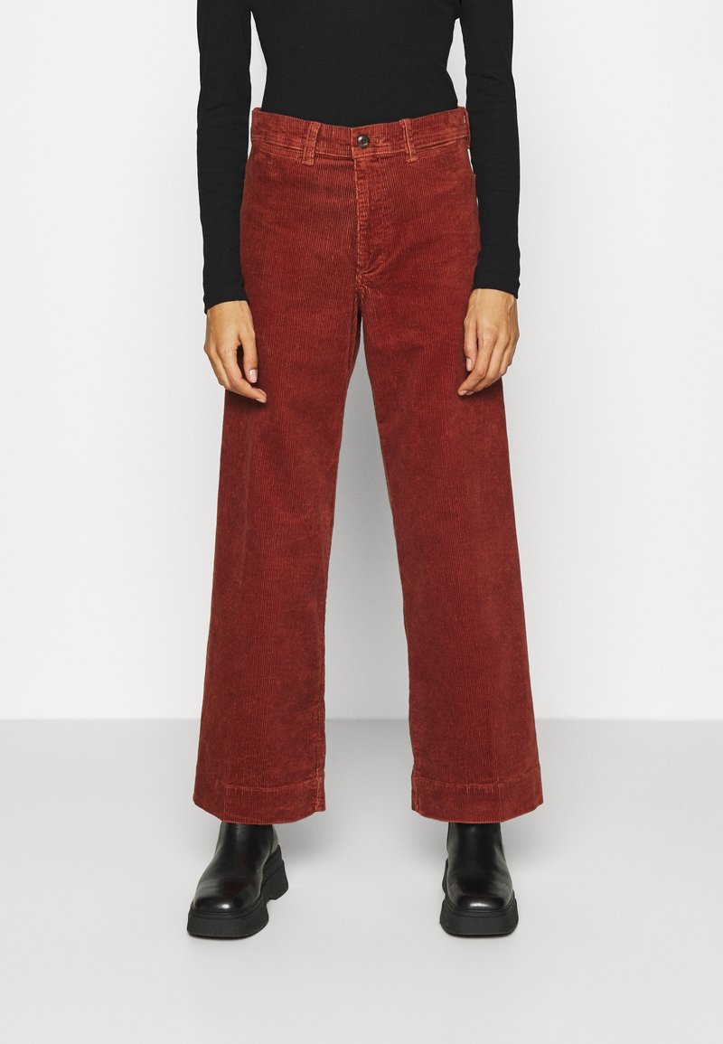 Personne portant un pantalon en velours côtelé large couleur rouille, un haut noir à manches longues et des bottines noires, debout devant un fond blanc.