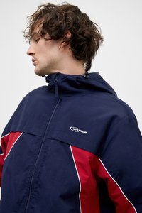 Αντιανεμικό μπουφάν σε ναυτικό μπλε και κόκκινο χρώμα με ψηλό γιακά, λευκή ρίγα, μπροστινό φερμουάρ και λογότυπο στο στήθος. Ελαφρύ, αθλητικό σχέδιο.