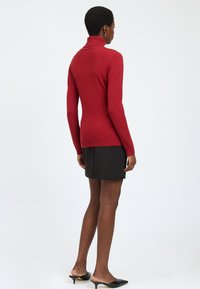 Maglione a collo alto rosso in tessuto a coste, design aderente, maniche lunghe, abbinato a una gonna corta nera, indossato con tacchi aperti.
