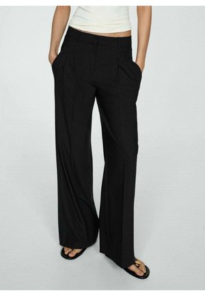 Kobieta ubrana w czarne spodnie z wysokim stanem i szerokimi nogawkami z plisami z przodu, z rękami w kieszeniach, oraz czarne sandały ze złotym zdobieniem.