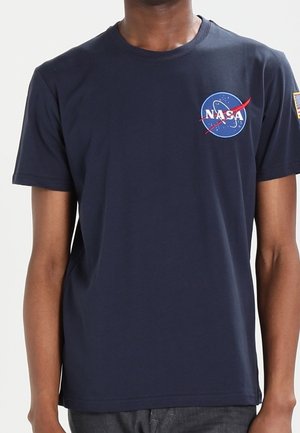 Person wearing a navy blue short-sleeve T-shirt with a NASA logo on the left chest and an American flag patch on the right sleeve.