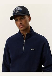 Navy zip-up pullover with a high collar and small logo patch; worn with a black cap featuring white and blue text design.