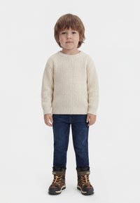 Jeune enfant debout, portant un pull en maille crème, un jean bleu foncé et des bottes d'hiver camouflage doublées en polaire.