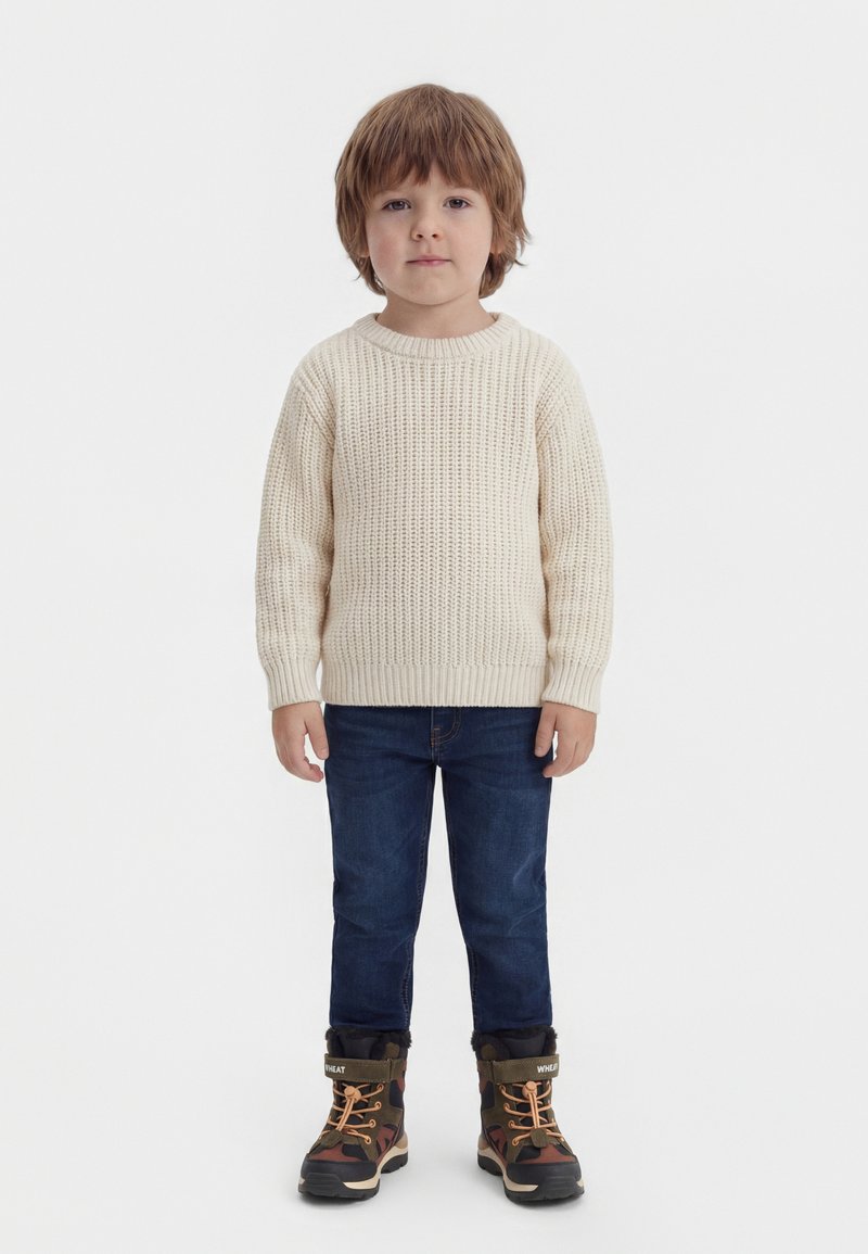 Jeune enfant debout, portant un pull en maille crème, un jean bleu foncé et des bottes d'hiver camouflage doublées en polaire.